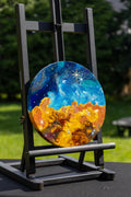 A round canvas painting displayed on a black easel outdoors. The artwork depicts a starforming region in blue and white with a glowing orange and yellow landscape below.