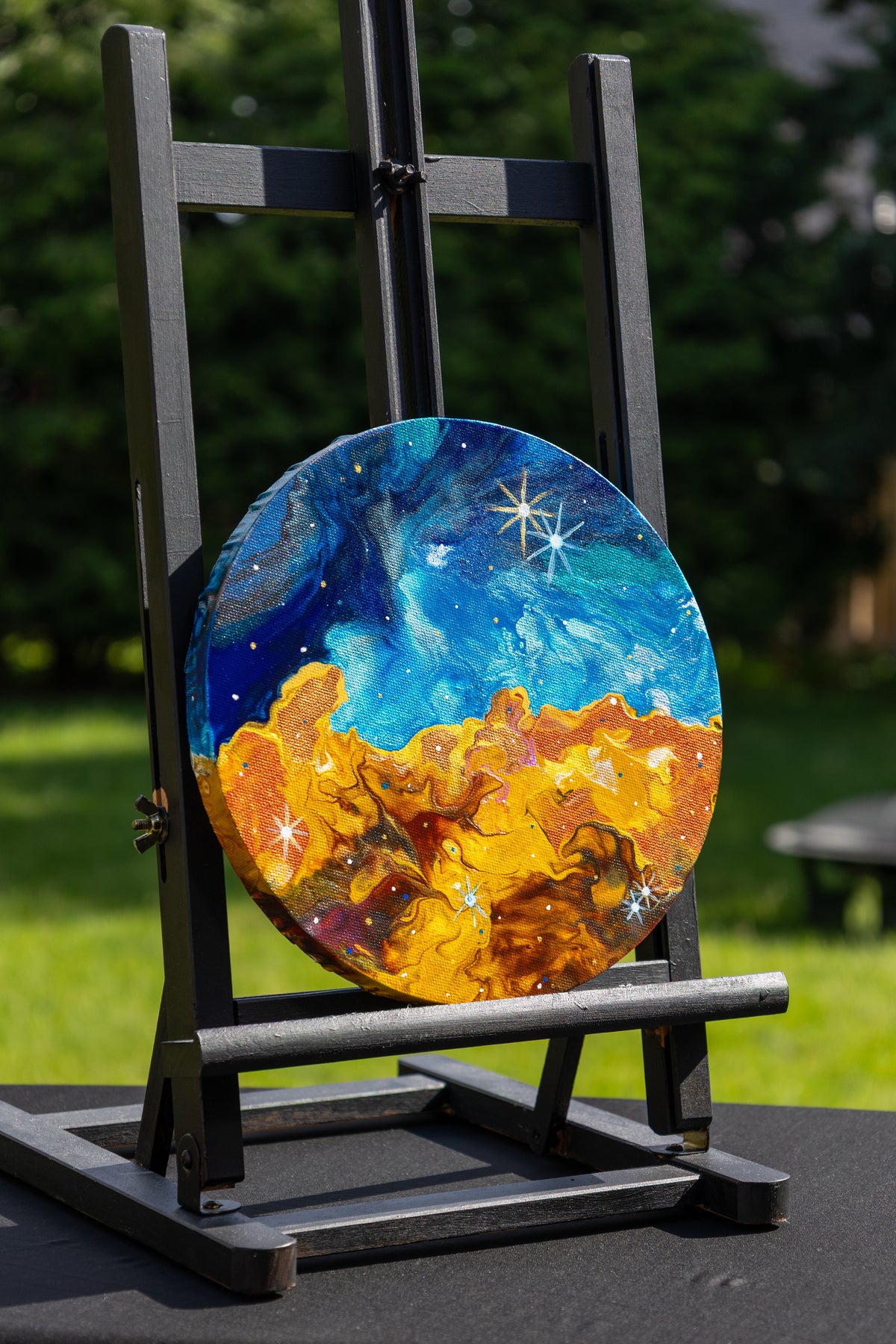 A round canvas painting displayed on a black easel outdoors. The artwork depicts a starforming region in blue and white with a glowing orange and yellow landscape below.
