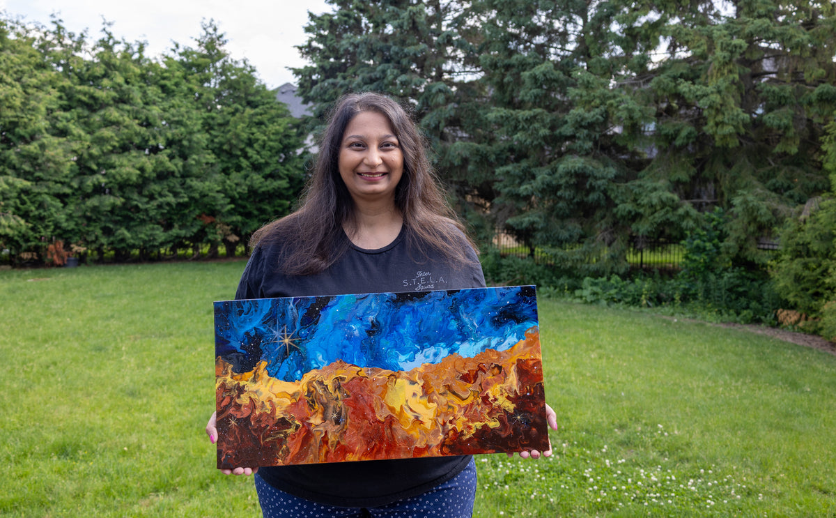 Artist Parshati Patel is standing in a grassy backyard holding up the Cosmicc Cliffs painting with blue, orange, and yellow tones.