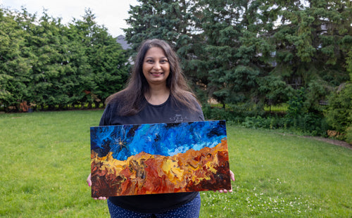 Artist Parshati Patel is standing in a grassy backyard holding up the Cosmicc Cliffs painting with blue, orange, and yellow tones.