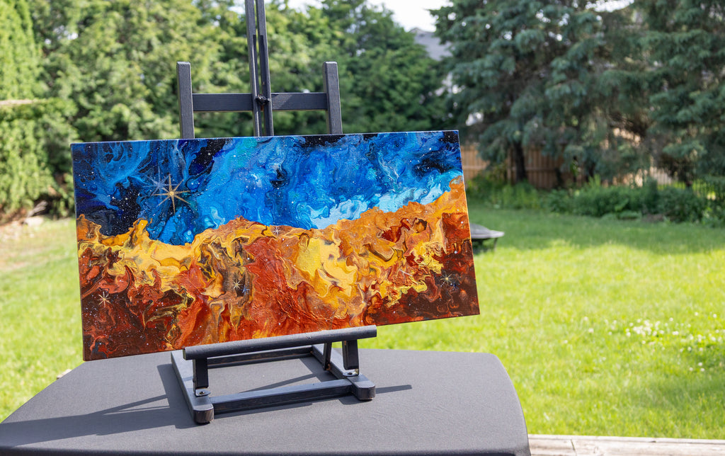 A space painting with a deep blue sky-like top half and fiery orange, yellow, and brown textured lower half, displayed on a black easel in an outdoor backyard setting with green grass and trees.