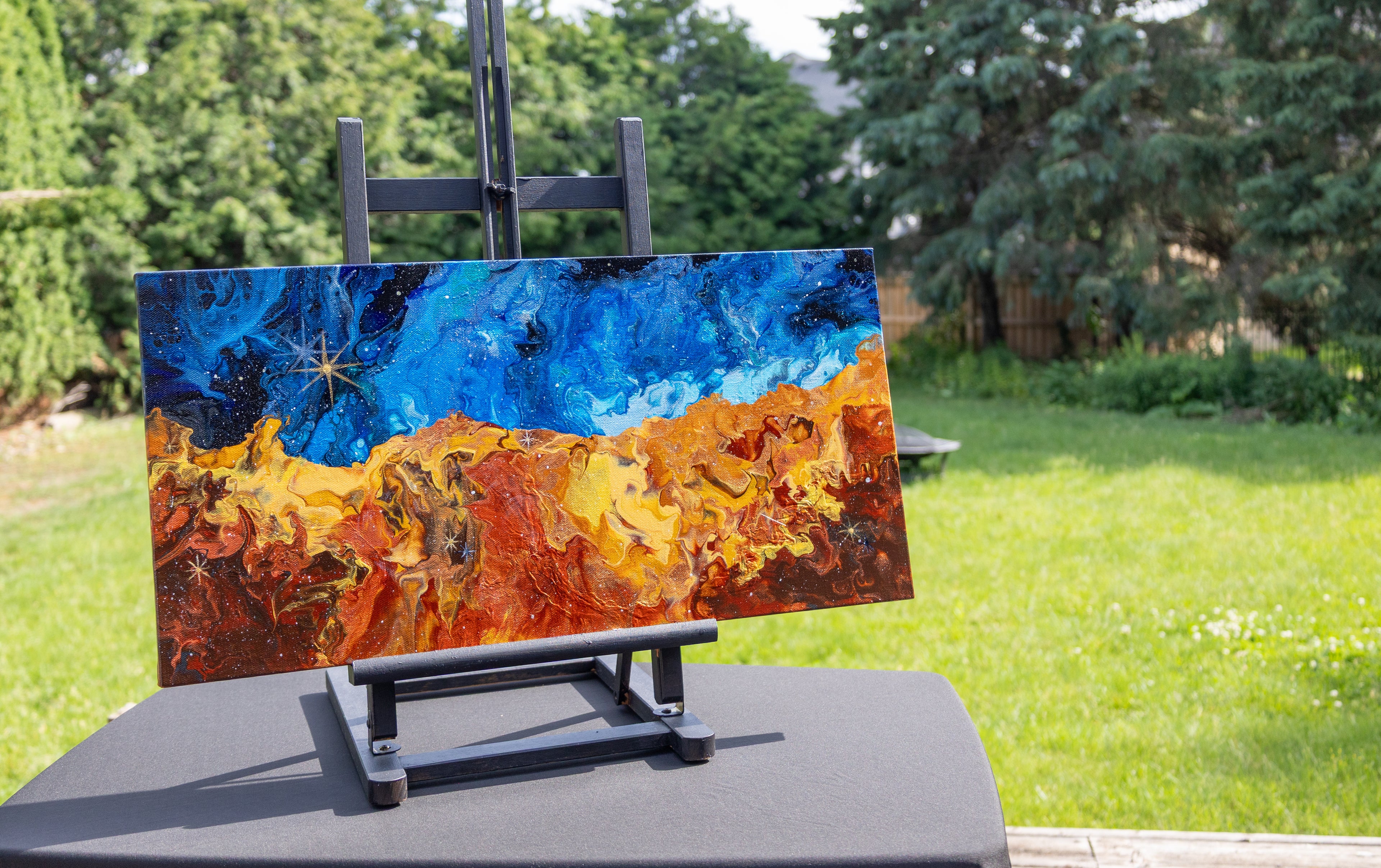 A space painting with a deep blue sky-like top half and fiery orange, yellow, and brown textured lower half, displayed on a black easel in an outdoor backyard setting with green grass and trees.