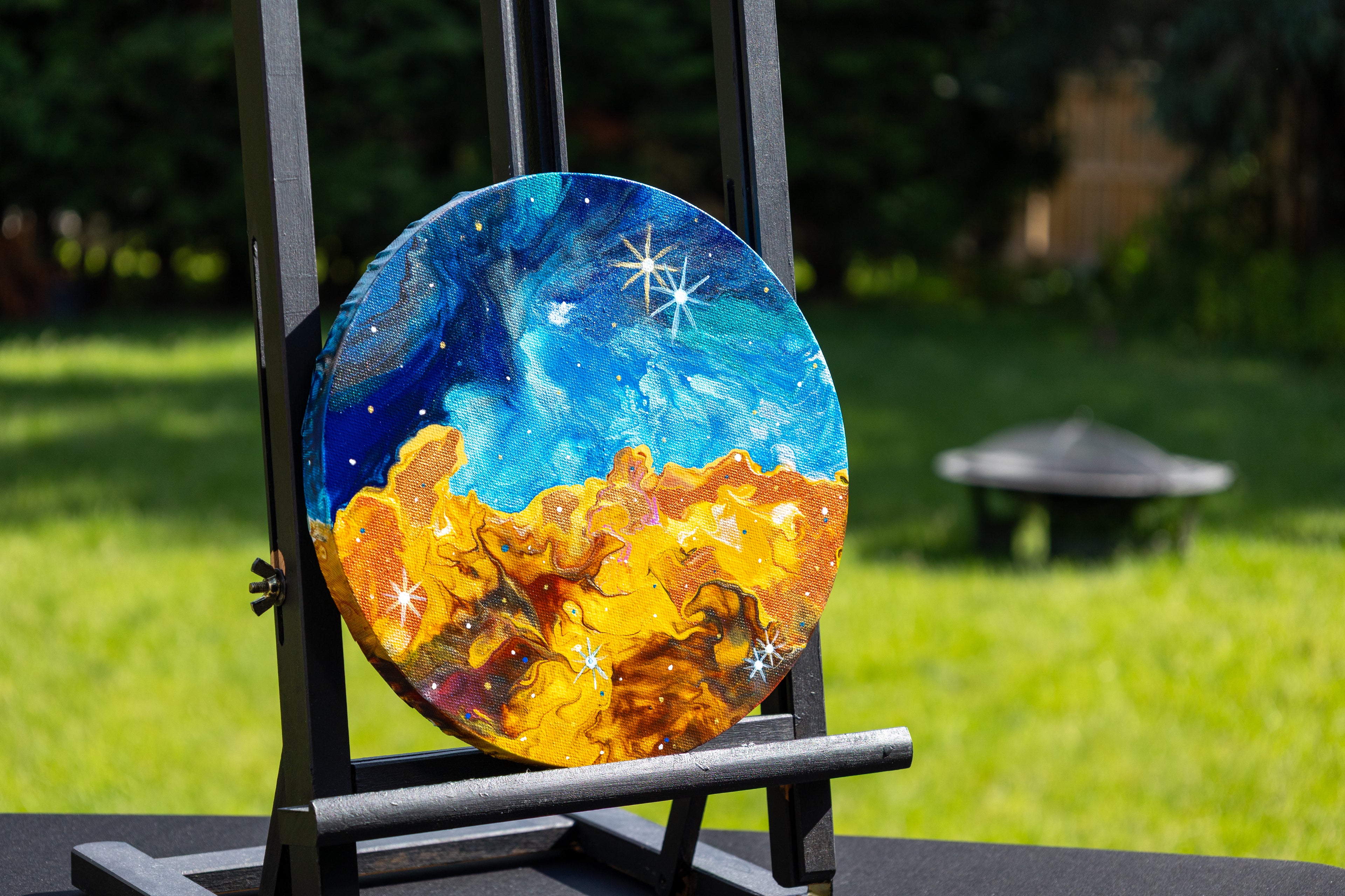 A round canvas painting displayed on a black easel outdoors. The artwork depicts a starforming region in blue and white with a glowing orange and yellow landscape below.