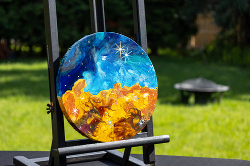 A round canvas painting displayed on a black easel outdoors. The artwork depicts a starforming region in blue and white with a glowing orange and yellow landscape below.