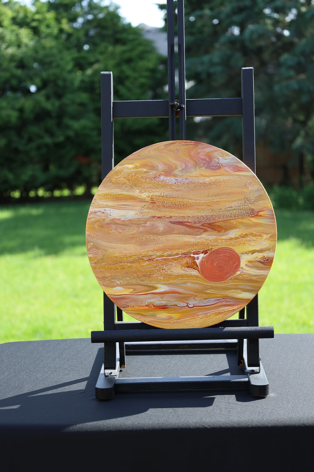 A round wooden plaque featuring a Jupiter-like pattern in orange, yellow, and brown tones, displayed on a black easel stand outdoors on a black table with a blurred green lawn and trees in the background.