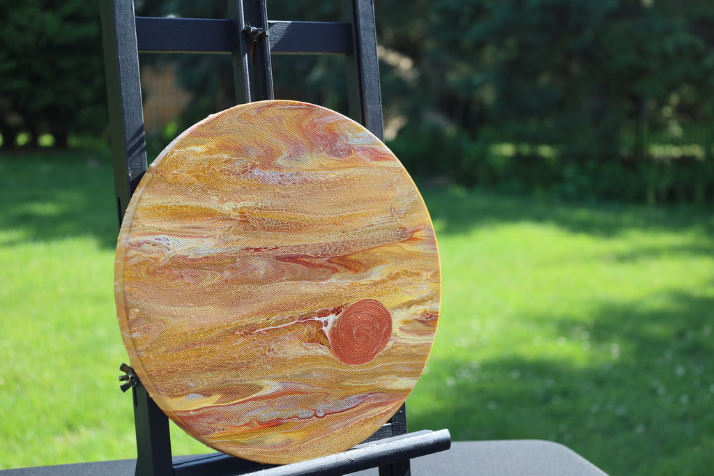 A round canvas painting featuring Jupiter  in shades of orange, gold, and brown, displayed on a black easel outdoors against a blurred green lawn background.