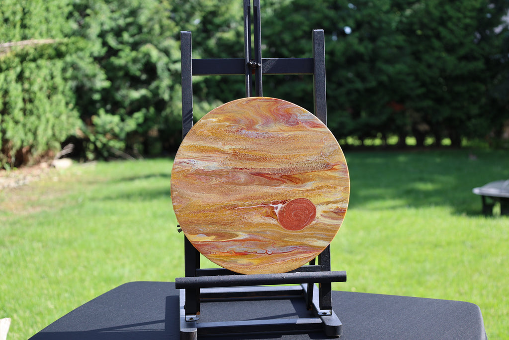 A round canvas featuring a Jupiter  in orange, brown, and cream, displayed on a black easel stand in an outdoor backyard setting with green grass and trees.