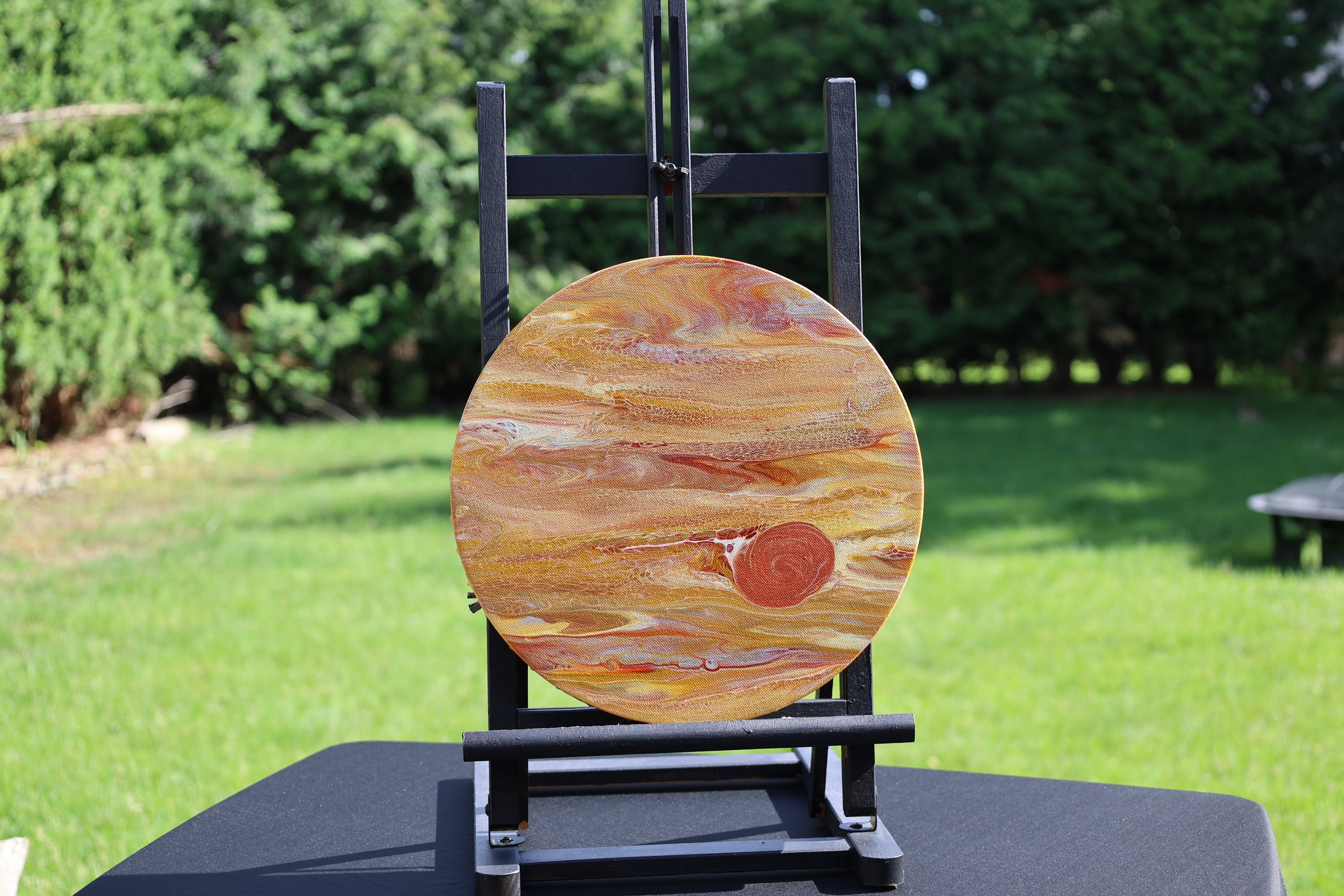 A round canvas featuring a Jupiter  in orange, brown, and cream, displayed on a black easel stand in an outdoor backyard setting with green grass and trees.