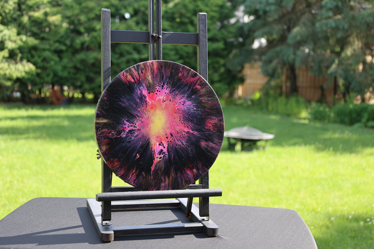 A round space-themed art piece with a colorful supernova design in purple, pink, orange, and black, displayed on a black metal stand in an outdoor backyard setting with a blurred green lawn and trees.