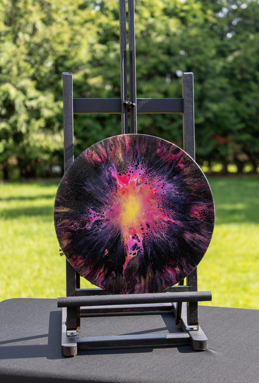 A round abstract art print featuring a vibrant supernova design in pink, purple, black, and yellow, displayed on a black metal easel in an outdoor garden setting.