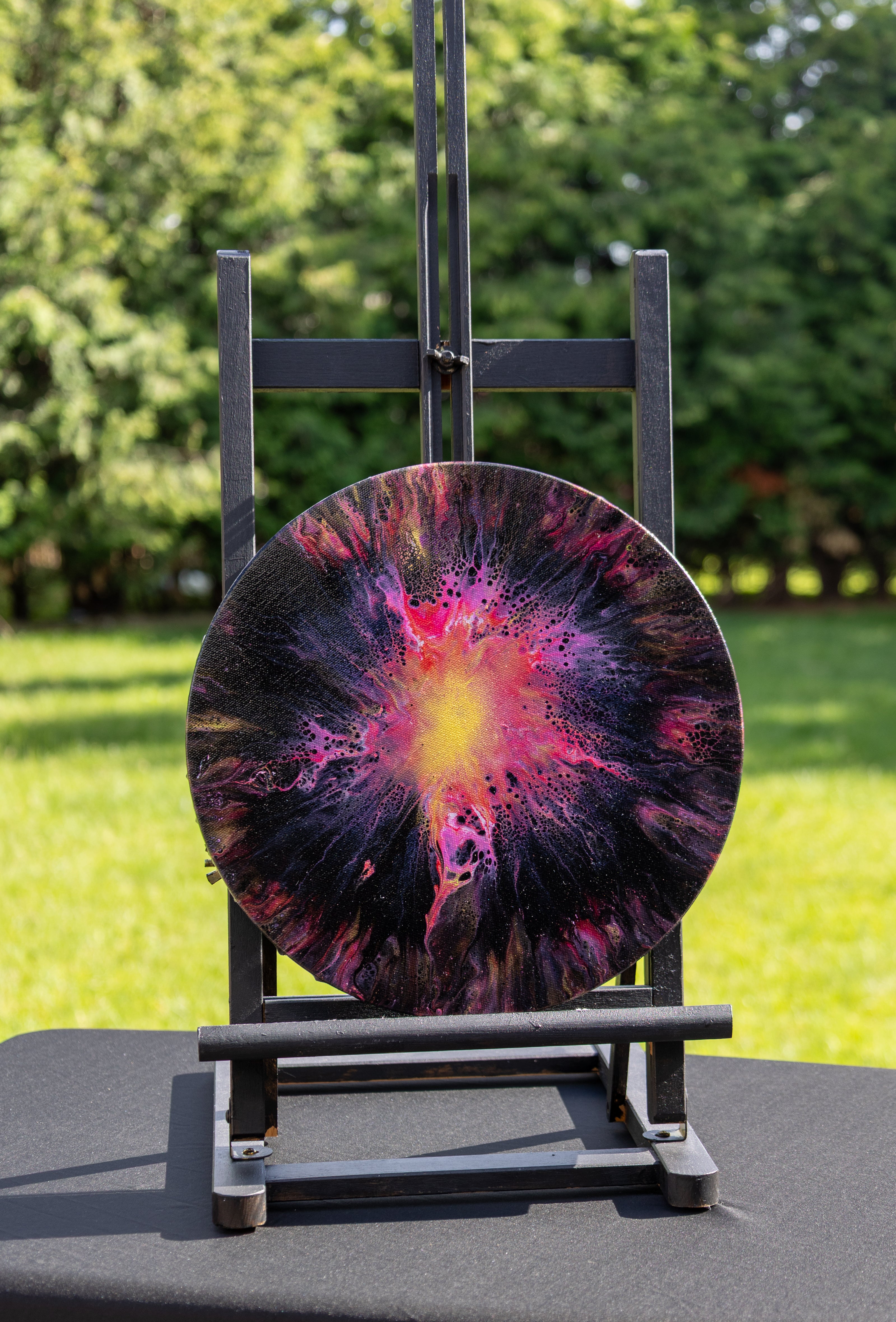 A round abstract art print featuring a vibrant supernova design in pink, purple, black, and yellow, displayed on a black metal easel in an outdoor garden setting.
